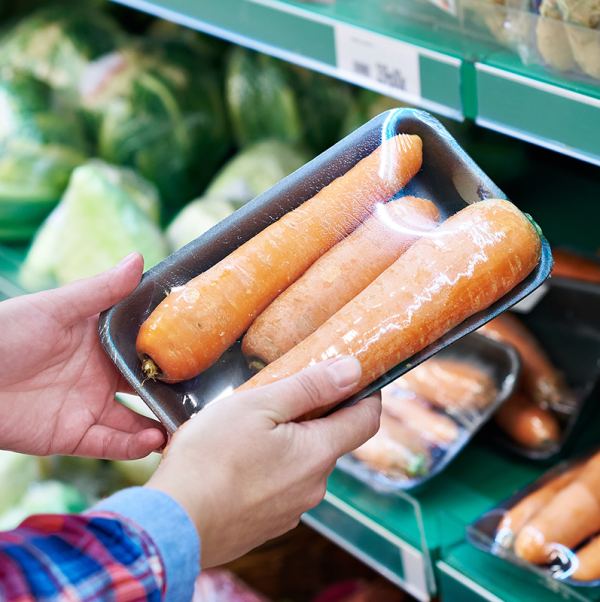 Un cliente en una tienda de comestibles mantiene una bandeja negra con tres zanahorias envueltas en envoltura retráctil